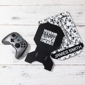 May include: A gray video game controller rests beside a black stand with the words "HUSBAND DADDY GAMER HERO" in white. A white and black patterned stand with the name "JAMES SMITH" is also present.