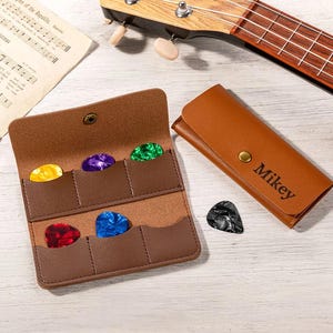 May include: A brown leather guitar pick holder with six colorful picks and a matching case with the name "Mikey" engraved. A ukulele is visible in the background, along with sheet music.