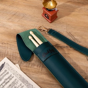 May include: A teal leather drumstick case with a green felt interior holds two wooden drumsticks. The case has a strap and a gold-colored clasp. The word "Oakley" is embossed on the case.