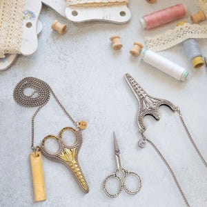 Può includere: Una vista dall'alto mostra accessori per cucire e gioielli. Due ciondoli a forma di forbici sono appesi a catene, uno oro e argento, l'altro argento. Un paio di piccole forbici d'argento sono vicine. Rocchetti di filo e pizzo sono sparsi intorno alle forbici.