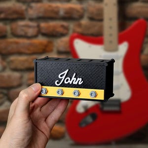Może przedstawiać: Czarny miniaturowy wzmacniacz z napisem "John" w białym skrypcie. Wzmacniacz ma żółty pasek ze srebrnymi pokrętłami. Czerwona gitara jest rozmyta w tle.