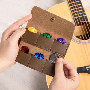 Könnte beinhalten: Ein brauner Leder-Gitarrenplektrumhalter mit sechs farbigen Plektren. Der Halter ist geöffnet und zeigt die Plektren in einzelnen Fächern. Im Hintergrund ist eine Gitarre zu sehen. Die Plektren sind gelb, grün, lila, rot, blau und grau.