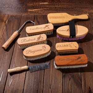 Op de afbeelding: Een verzameling houten verzorgingsgereedschappen voor paarden, waaronder borstels, een kam en een hoevenkrabber. De borstels zijn gegraveerd met namen en paardgerelateerde ontwerpen. De gereedschappen liggen op een donker houten oppervlak.
