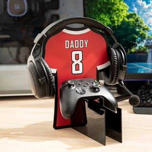 May include: A black gaming headset sits on a red jersey-shaped stand with the word "DADDY" and the number "8" printed on it. A black game controller is positioned below the jersey. The stand is on a wooden surface.