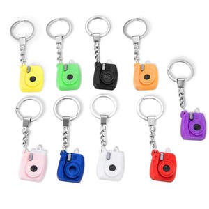 Op de afbeelding: Een verzameling camera-vormige sleutelhangers in verschillende kleuren. De sleutelhangers zijn geel, groen, zwart, oranje, roze, blauw, wit, rood en paars. Elke sleutelhanger heeft een zilveren metalen ring en ketting.