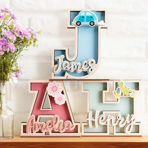 Può includere: Salvadanai a forma di lettera in legno con nomi. La lettera "J" è azzurra con un'auto, la lettera "A" è rosa con fiori e la lettera "H" è verde chiaro con un dinosauro. I nomi "James", "Amelia" e "Henry" sono scritti sulle lettere.