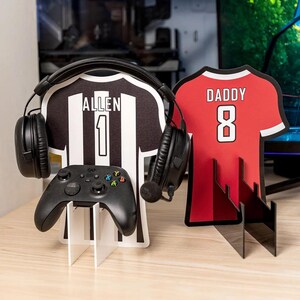 Puede incluir: Un soporte en forma de camiseta en blanco y negro con el nombre "ALLEN" y el número "1" impresos, sosteniendo un mando de Xbox negro y unos auriculares negros. También hay un soporte en forma de camiseta roja con la palabra "DADDY" y el número "8".