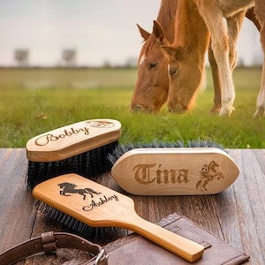 May include: Three wooden horse grooming brushes with personalized names and a horse silhouette. The brushes have black bristles and are displayed on a wooden surface. Two horses graze in the background.