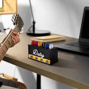 Könnte beinhalten: Ein schwarzer Gitarrenplektrumhalter in Form eines Verstärkers mit dem Namen "Daisy" in weißer Schrift. Er enthält mehrere bunte Gitarrenplektren. Der Halter hat gelbe Akzente und Knöpfe und steht auf einem Schreibtisch neben einem Laptop.