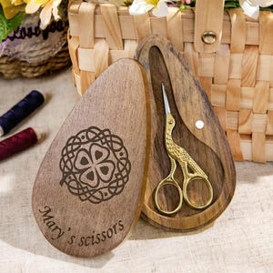 Pode incluir: Uma caixa de madeira com um design decorativo de trevo e o texto "Mary's scissors" contém um par de tesouras douradas. As tesouras têm um design semelhante a um pássaro. A caixa está aberta, revelando as tesouras no interior.