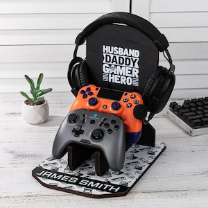 May include: A black wooden gaming headset stand with the text "HUSBAND DADDY GAMER HERO". It holds an orange and a gray game controller. The base has a pattern of game controllers and the name "JAMES SMITH".
