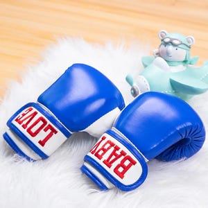 Può includere: Un paio di guantoni da boxe blu con bordini bianchi e testo ricamato in rosso. Un guanto dice "BABY" e l'altro dice "LOVE".