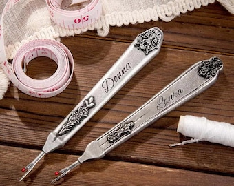 May include: Two silver seam rippers with ornate floral designs and the names "Donna" and "Laura" engraved on them. A pink and white measuring tape and a spool of white thread are also present.