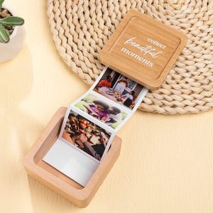 以下が含まれることがあります： 「collect beautiful moments」の文字が蓋に刻まれた木製のフォトボックス。ボックスは開いており、一連の小さな写真が写っています。ボックスは明るい色の木でできており、織り込まれたマットの上に置かれています。