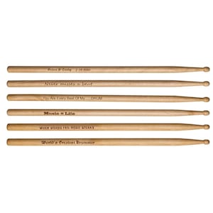 Puede incluir: Seis baquetas de batería de madera con texto grabado. El texto incluye frases como "Never misses a beat" y "Music = Life". Un par está personalizado con un nombre y una fecha. Las baquetas son de color marrón claro.