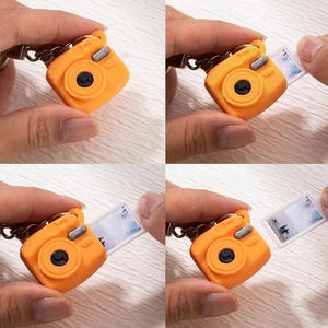 Könnte beinhalten: Ein orangefarbener Miniatur-Kamera-Schlüsselanhänger mit schwarzem Objektiv und silbernen Akzenten. Ein kleines Foto gleitet aus der Kamera und zeigt eine Landschaft. Der Schlüsselanhänger wird von einer Hand gehalten.