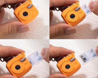 May include: A miniature orange camera keychain with a black lens and silver details. A small photo slides out of the camera, showing a landscape scene. The keychain is held by a person's hand.