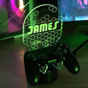 May include: A black gaming controller with a green illuminated acrylic display. The display features the name "JAMES" in bold, green letters, set against a honeycomb pattern. The controller is resting on a black base.