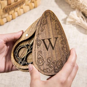 Può includere: Una scatola di legno a forma di goccia con un design ornato e la lettera "W" incisa sul coperchio. All'interno, un paio di forbici decorative in stile antico. La scatola è aperta, rivelando le forbici e i dettagli del legno.