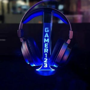 Puede incluir: Auriculares plateados y negros sobre un soporte iluminado en azul. El soporte muestra la palabra "GAMER123" en blanco. El soporte y los auriculares están iluminados en azul.
