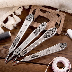 May include: Four silver seam rippers with floral detailing and names: Donna, Sally, Elaine, and Laura. A wooden sewing machine cutout and measuring tape are also visible.