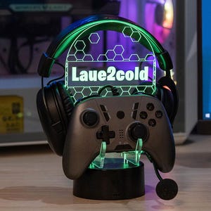 May include: A gaming accessory stand with a controller and headset. The stand has a black base and a clear acrylic top with the text "Laue2cold" illuminated in white. The headset is black with green accents.