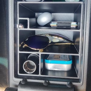 May include: A black plastic car center console organizer insert with multiple compartments. The organizer holds various items including a pair of sunglasses, a metal tin, a small wallet, a round gray object, a cylindrical metal tool, and a pen.