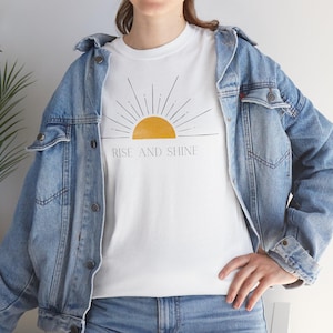 Op de afbeelding: Wit T-shirt met een gele zon-afbeelding en de tekst "RISE AND SHINE". Het shirt wordt gecombineerd met een blauw spijkerjasje en een jeans. Het ontwerp is eenvoudig en de tekst is in een strak lettertype.