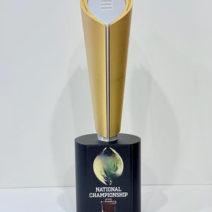 May include: A gold-colored trophy with a black base. The trophy has a unique shape with a white top and the words "NATIONAL CHAMPIONSHIP" and the year "2025" on the base.