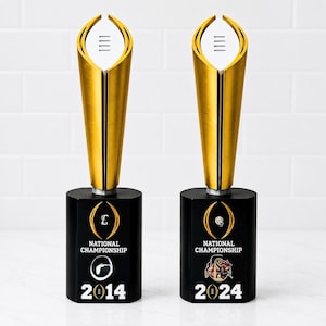 May include: Two gold-colored football trophies with a tapered top and a black cylindrical base. Each base features the text National Championship, a logo, and the years 2014 and 2024 respectively, with a small football icon above the year.