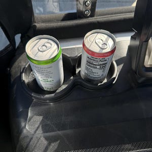 May include: Two aluminum beverage cans in a golf cart cup holder. One can is white with green accents and text that reads "A Refreshing Spiked Sparkling Water with Natural Flavors & 100 Calories". The other can is red and white with nutrition facts.