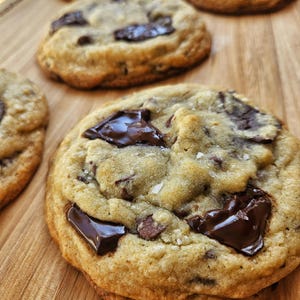 Puede incluir: Primer plano de una galleta de chocolate con trozos grandes de chocolate sobre una superficie de madera. La galleta es de color marrón dorado y tiene una textura ligeramente crujiente.