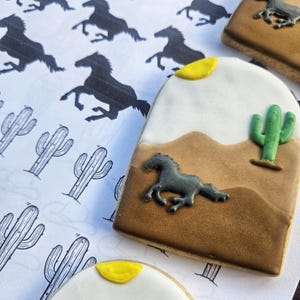 Puede incluir: Dos galletas de az&uacute;car decoradas con una escena del desierto. Las galletas son marrones, blancas y amarillas con una silueta negra de un caballo corriendo. Una galleta tiene un cactus verde.