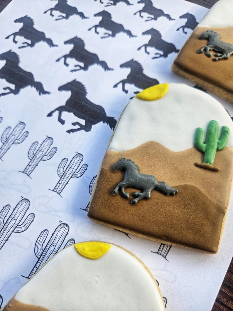 Puede incluir: Dos galletas de az&uacute;car decoradas con una escena del desierto. Las galletas son marrones, blancas y amarillas con una silueta negra de un caballo corriendo. Una galleta tiene un cactus verde.