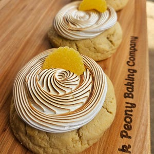 Puede incluir: Primer plano de tres galletas de merengue de limón con una cobertura de caramelo amarillo sobre una tabla de madera con el texto "Pink Peony Baking Company" grabado en la tabla.