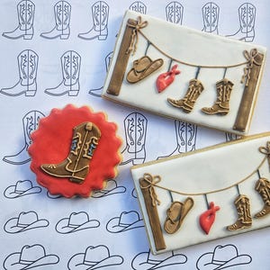 Puede incluir: Tres galletas de az&uacute;car decoradas. Una galleta es roja con una bota de vaquero marr&oacute;n. Las otras dos galletas son blancas con una cuerda marr&oacute;n y botas de vaquero marrones, sombreros y pa&ntilde;uelos colgando de la cuerda.