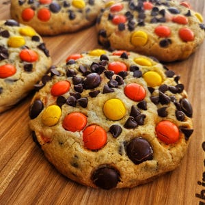 Puede incluir: Primer plano de varias galletas recién horneadas sobre una tabla de cortar de madera. Las galletas son de color marrón dorado y están salpicadas de chispas de chocolate, caramelos amarillos y naranjas. Las galletas son un manjar.