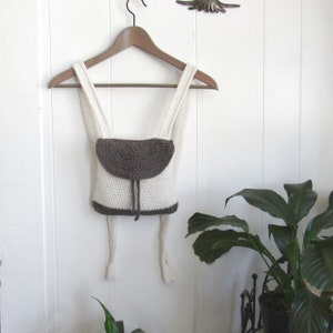 May include: A small white crocheted backpack with a brown crocheted flap. The backpack is hanging on a wooden hanger against a white wall.