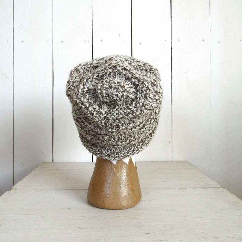 May include: A brown and white knitted beanie hat on a wooden display stand. The hat has a textured, chunky knit pattern.