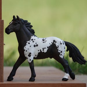 May include: A toy horse figurine with a black head, neck, and legs, featuring a white patch on its midsection covered in black spots. The horse has a black mane and tail, a white blaze on its face, and a white marking on its rear left hoof.