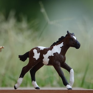 May include: A small toy horse figurine featuring a dark brown coat with irregular white patches across its body, a black mane and tail, and white markings on its lower legs. The horse is depicted in a trotting pose with one front leg raised.