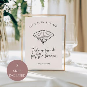 May include: A framed sign with a gold border, displaying the words "LOVE IS IN THE AIR" above a fan illustration. The text "Take a fan & feel the breeze" and "SARAH & MIKE" are below. A circular graphic indicates "2 SIZES INCLUDED".