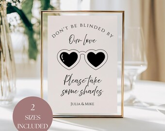 May include: A framed sign with gold trim features the text "DON'T BE BLINDED BY Our love" above heart-shaped sunglasses and the phrase "Please take some shades." The sign includes the names "JULIA & MIKE" and a circle that says "2 SIZES INCLUDED."