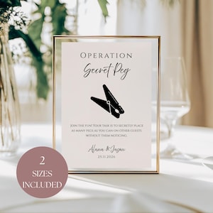 Puede incluir: Un letrero enmarcado con el texto "OPERATION Secret Peg" y un gráfico de pinza de ropa negra. El letrero incluye instrucciones y los nombres "Alana & Jason" con una fecha. Una etiqueta circular indica "2 TAMAÑOS INCLUIDOS".