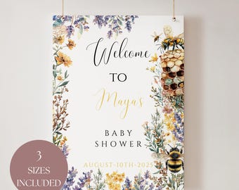 Op de afbeelding: Een babyborrelbord met bloemen- en bijenillustraties. Op het bord staat "Welkom bij Maya's Baby Shower" met de datum 10 augustus 2025. Het bord is versierd met lavendel, gele en oranje bloemen en een honingraat.