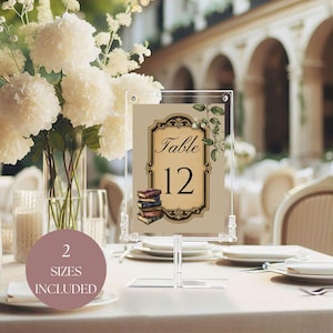 May include: Clear acrylic table number card with a vintage design. The card features the number 12 in a decorative frame with a stack of books and floral accents. The text "Table" is also visible. A round sticker with the text "2 SIZES INCLUDED" is in the foreground.