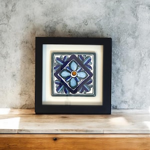 Puede incluir: Un azulejo de cerámica enmarcado con un diseño floral azul y blanco. El azulejo está colocado sobre un fondo gris.