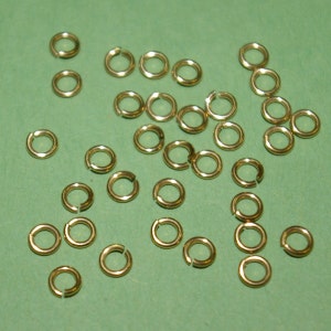 May include: A large quantity of gold jump rings, scattered on a green surface. The jump rings are small, circular, and made of metal.