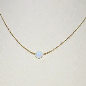 May include: A delicate gold chain necklace featuring a single, round, iridescent opal pendant. The opal displays a play of colors, including white, blue, and green. The necklace is displayed against a neutral background.