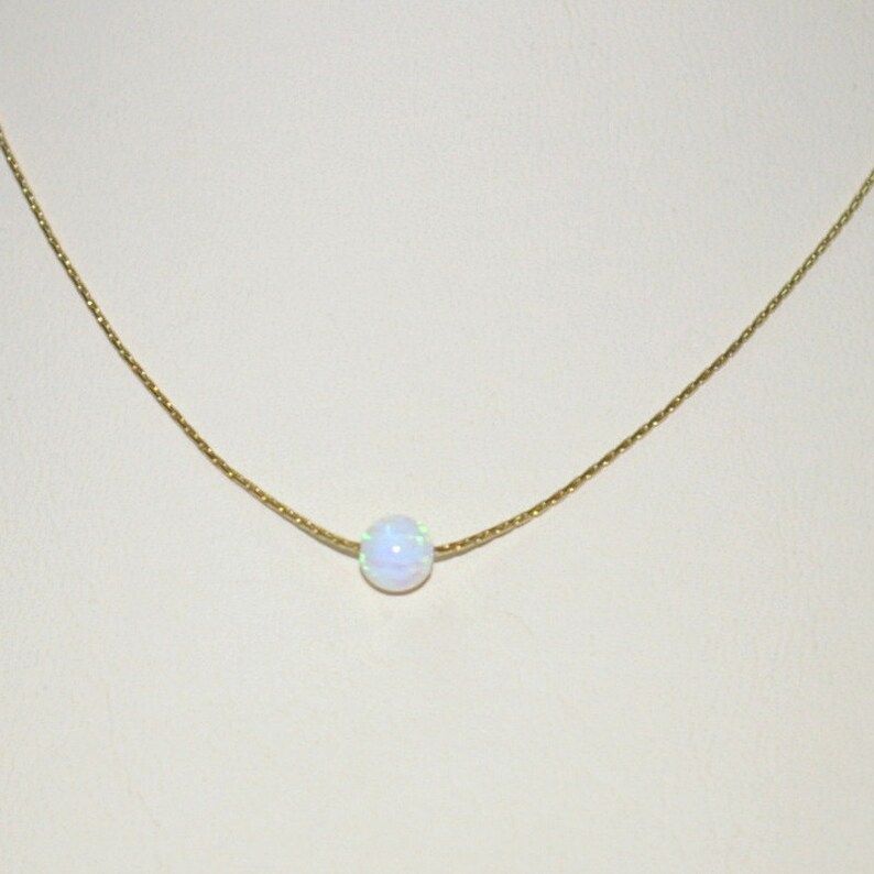 May include: A delicate gold chain necklace featuring a single, round, iridescent opal pendant. The opal displays a play of colors, including white, blue, and green. The necklace is displayed against a neutral background.